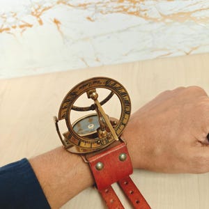 May include: A vintage brass sundial watch with a red leather strap. The sundial features Roman numerals and the words "West London" engraved on the outer ring. The watch is designed to be worn on the wrist.