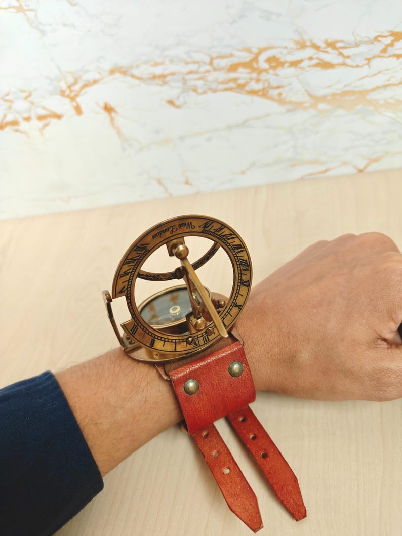 May include: A vintage brass sundial watch with a red leather strap. The sundial features Roman numerals and the words "West London" engraved on the outer ring. The watch is designed to be worn on the wrist.