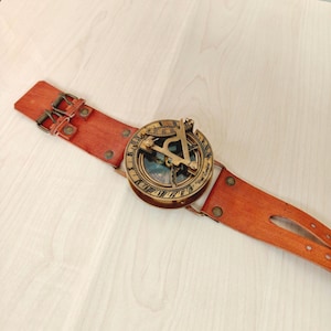 May include: A vintage-style sundial compass watch with a brown leather strap. The compass is round with a brass finish and intricate detailing. The leather strap has a buckle closure and is riveted.