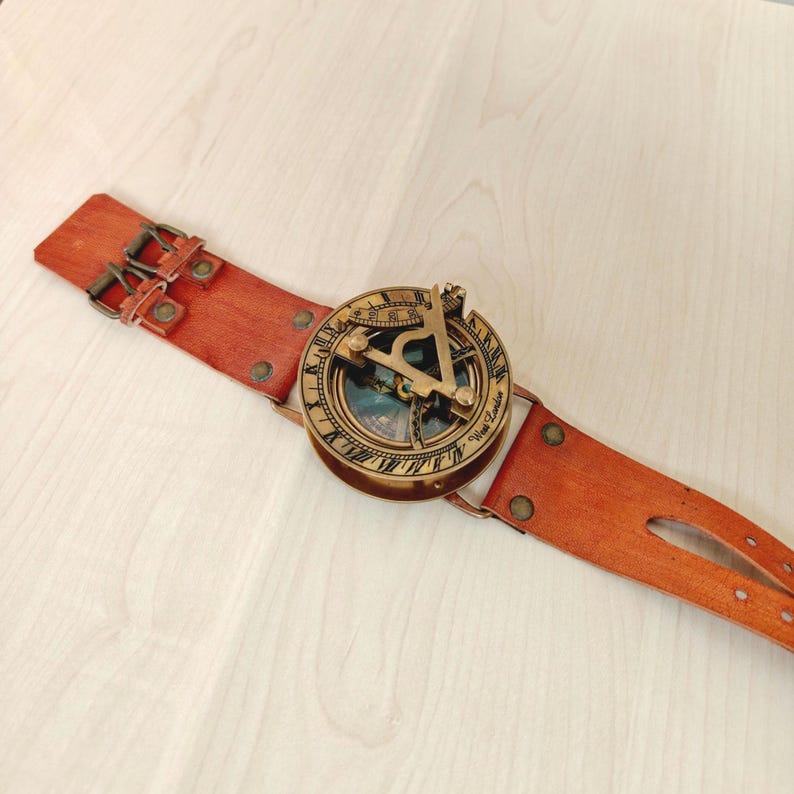 May include: A vintage-style sundial compass watch with a brown leather strap. The compass is round with a brass finish and intricate detailing. The leather strap has a buckle closure and is riveted.