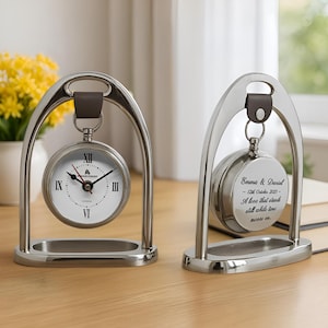 May include: Two silver desk clocks with a distinctive arched frame. One clock face shows Roman numerals and the text "Diamond Street London". The other clock has the inscription "Emma & Daniel - 12th October 2025 - A love that stands still while time moves on."