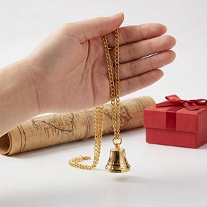 Puede incluir: Un collar con colgante de campana dorada con una cadena es sostenido por una mano. Un mapa enrollado y una caja de regalo roja con un lazo están en el fondo. El colgante de campana mide aproximadamente 2,5 cm de altura.