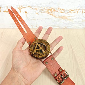 May include: A vintage-style sundial watch with a brass face and a reddish-brown leather strap. The watch features a compass design and is secured with two buckles. The leather strap measures approximately 10 inches in length.
