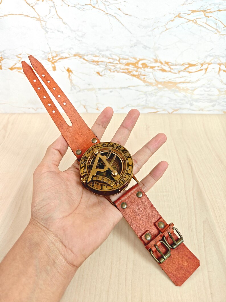 May include: A vintage-style sundial watch with a brass face and a reddish-brown leather strap. The watch features a compass design and is secured with two buckles. The leather strap measures approximately 10 inches in length.