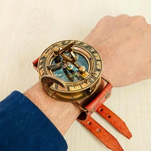 May include: A vintage brass sundial watch with a leather strap. The watch face features Roman numerals and the text "West London." The watch has a compass and is attached to a red leather strap.