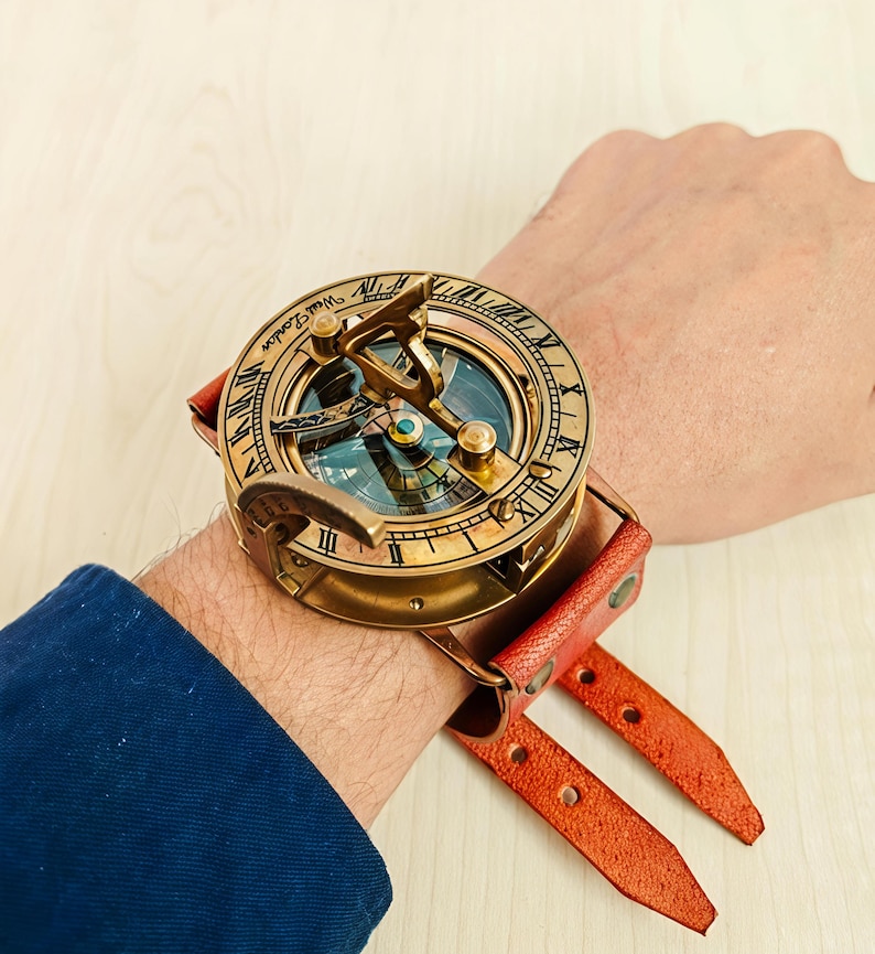 May include: A vintage brass sundial watch with a leather strap. The watch face features Roman numerals and the text "West London." The watch has a compass and is attached to a red leather strap.