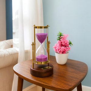May include: An ornate hourglass with a brass frame and a wooden base. The glass chamber is filled with purple sand. The hourglass sits on a wooden table, next to a small white pot with pink flowers. The background features a window and a light blue wall.