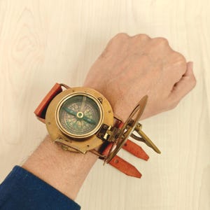 May include: A vintage brass compass watch with a leather strap. The compass face features a detailed directional dial with a green and gold color scheme. The watch has a hinged cover and is attached to a reddish-brown leather strap.