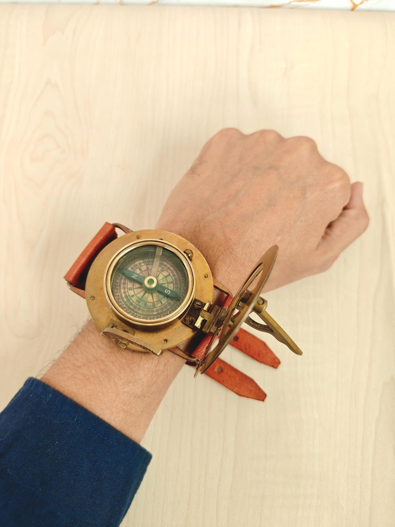 May include: A vintage brass compass watch with a leather strap. The compass face features a detailed directional dial with a green and gold color scheme. The watch has a hinged cover and is attached to a reddish-brown leather strap.