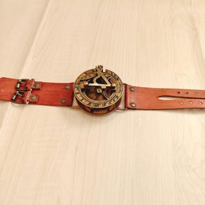 May include: A vintage-style sundial watch with a brown leather strap. The watch face is gold-colored with intricate markings and a triangular gnomon. The leather strap has two buckles and several adjustment holes.