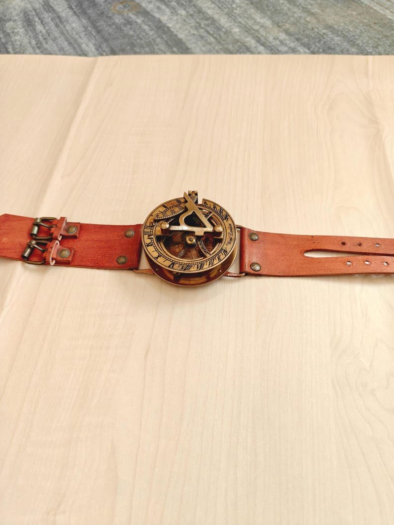 May include: A vintage-style sundial watch with a brown leather strap. The watch face is gold-colored with intricate markings and a triangular gnomon. The leather strap has two buckles and several adjustment holes.
