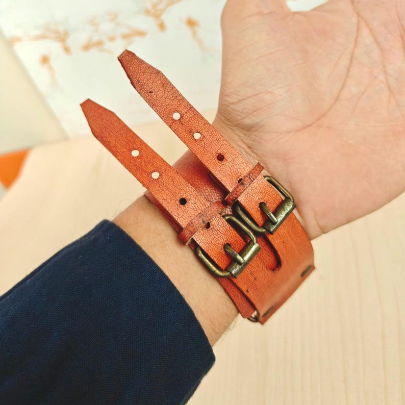 May include: A close-up of an orange leather wrist cuff with two straps and brass buckles. The cuff is worn on a person's wrist, with the straps buckled. The person is wearing a navy blue long-sleeve shirt.