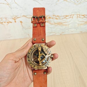 May include: A vintage-style brass sundial compass with a leather strap. The compass face features intricate markings and a gnomon. The leather strap is a reddish-brown color and has two buckles. The text "West London" is visible on the compass.