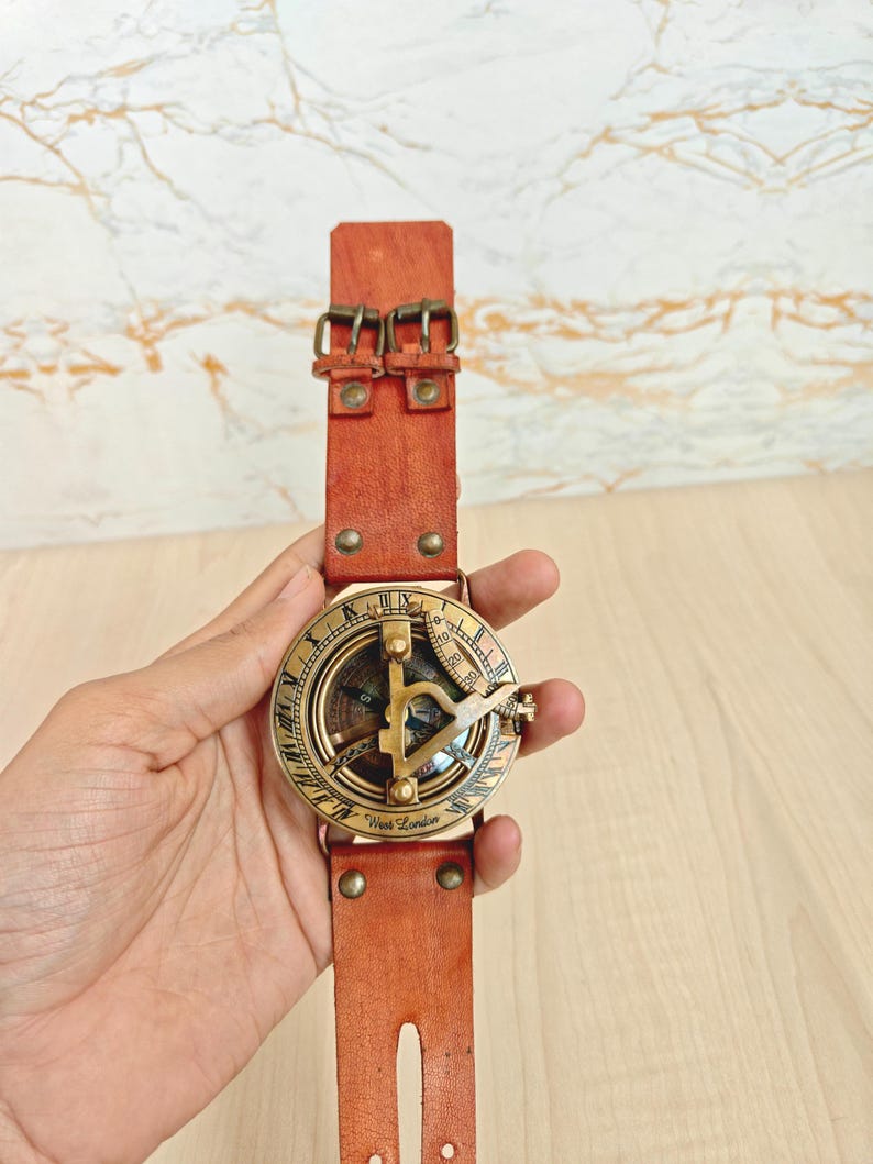 May include: A vintage-style brass sundial compass with a leather strap. The compass face features intricate markings and a gnomon. The leather strap is a reddish-brown color and has two buckles. The text "West London" is visible on the compass.