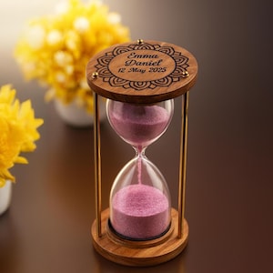 May include: A wooden hourglass with a decorative top and base, connected by gold-colored metal rods. The hourglass contains pink sand, with the text "Emma Daniel 12 May 2025" engraved on the top. The background features yellow flowers.