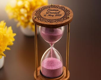 Può includere: Una clessidra in legno con parte superiore e base decorative, collegate da aste metalliche color oro. La clessidra contiene sabbia rosa, con il testo "Emma Daniel 12 May 2025" inciso sulla parte superiore. Fiori gialli sullo sfondo.