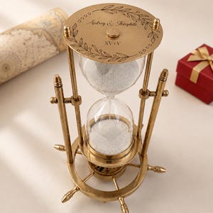 May include: A brass hourglass with a ship wheel base and white sand. The top plate is engraved with a leaf border, the text Audrey & Théophile, and the Roman numerals XV+V. The structure features vertical supports and a central glass bulb.