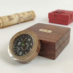 May include: A brass compass with the inscription "So you can always find your way back home. Love Anna." is displayed next to a wooden box with the initials "A.N." engraved on it. A rolled-up map and a red gift box are in the background.