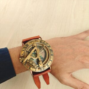 May include: A vintage brass sundial watch with a brown leather strap. The watch face features intricate detailing and a compass design. The watch is worn on the wrist, showcasing its antique aesthetic.