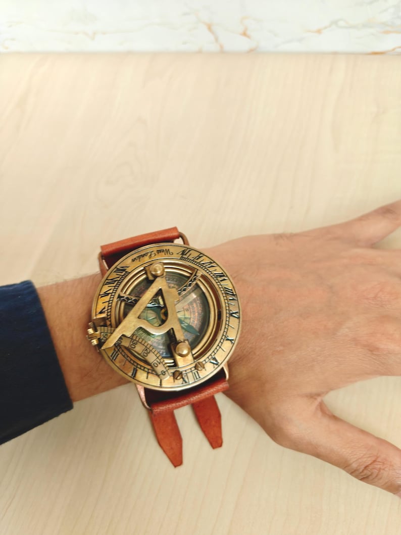 May include: A vintage brass sundial watch with a brown leather strap. The watch face features intricate detailing and a compass design. The watch is worn on the wrist, showcasing its antique aesthetic.
