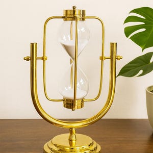 Puede incluir: Un reloj de arena dorado con una cámara de vidrio transparente llena de arena blanca. El reloj de arena tiene un marco de metal curvo decorativo y una base circular. El diseño es elegante y adecuado para la decoración del hogar.