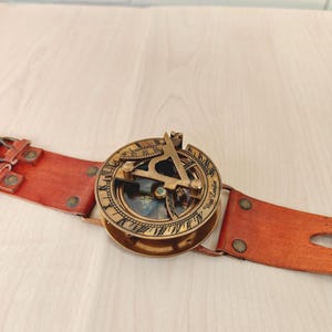 May include: A vintage brass sundial compass with a leather strap. The compass features intricate detailing and a glass-covered dial. The leather strap is a reddish-brown color and has metal rivets. The compass is a navigational tool.