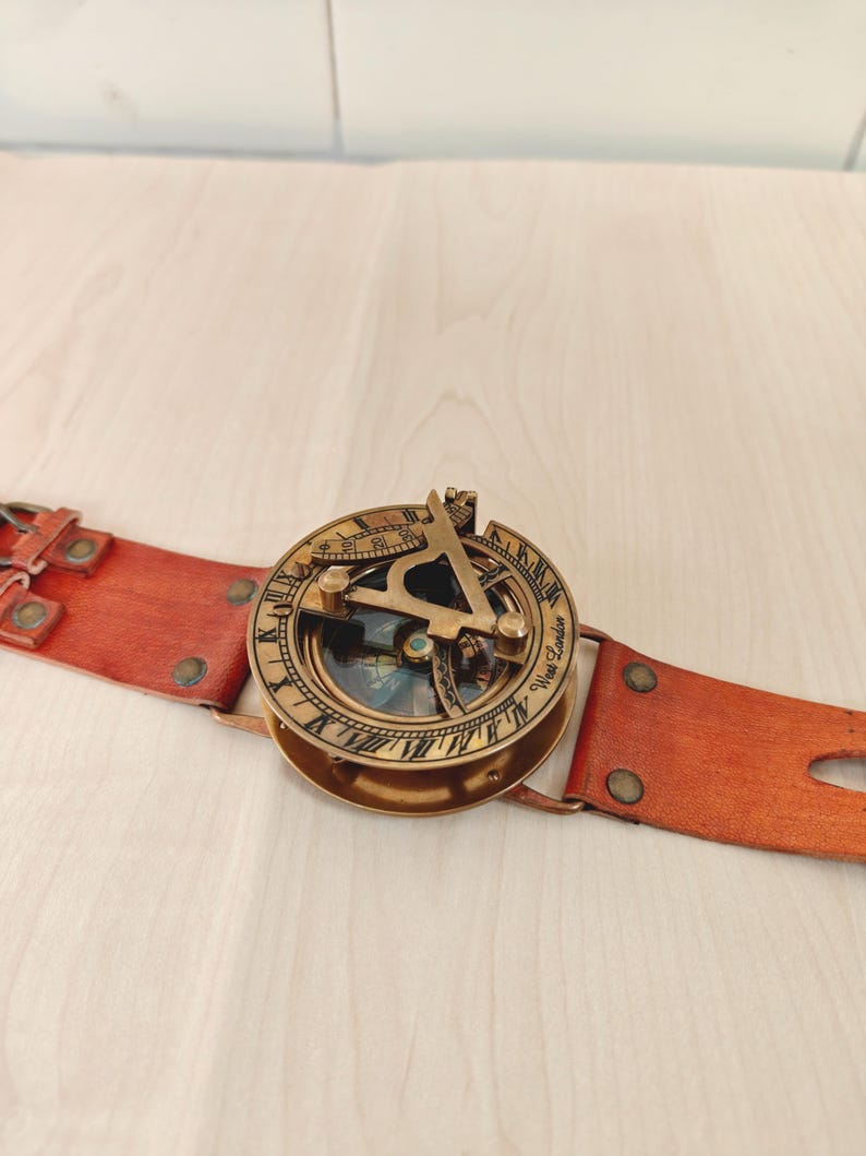 May include: A vintage brass sundial compass with a leather strap. The compass features intricate detailing and a glass-covered dial. The leather strap is a reddish-brown color and has metal rivets. The compass is a navigational tool.