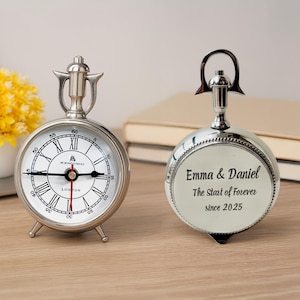 Puede incluir: Dos relojes decorativos plateados. El reloj de la izquierda tiene una esfera blanca con números romanos negros y el texto "49 Bond Street London". El reloj de la derecha tiene el texto "Emma & Daniel The Start of Forever since 2025".