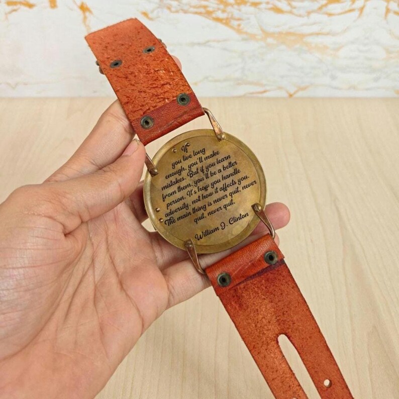May include: A vintage-style leather belt with a circular brass buckle. The buckle features an engraved quote by William J. Clinton. The belt is a reddish-brown color and has a distressed appearance.