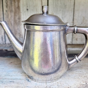 May include: A metallic silver-toned teapot featuring a rounded body, a curved spout, and a matching lid with a small knob handle. The teapot has a sturdy side handle attached with two distinct metal bands, showcasing a weathered, textured surface.