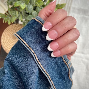 May include: Close-up of a hand with oval-shaped nails featuring a classic French manicure. The nails have a pale pink base with white tips. The hand is next to a blue denim jacket with tan stitching.