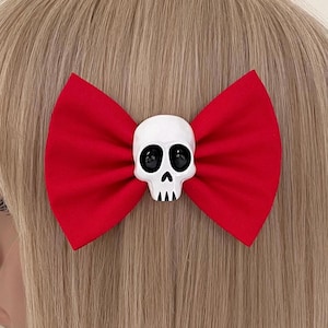 Puede incluir: Una pinza para el pelo con lazo de tela roja con un cráneo blanco en el centro. El cráneo tiene ojos negros y una boca pequeña. Un accesorio para el cabello divertido y espeluznante.