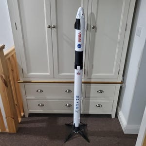 May include: A white and black model rocket with the words "SpaceX" and "NASA" printed on the side. The rocket is standing on a black tripod stand.