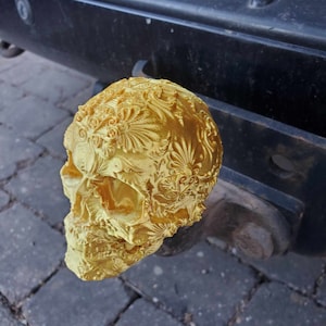 Könnte beinhalten: Eine goldfarbene Schädelskulptur mit aufwendigen floralen Details. Der Schädel ist aus einem glatten, glänzenden Material gefertigt und hat ein detailliertes Design.