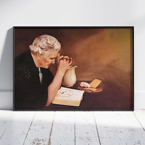 Op de afbeelding: Een vrouw in een zwarte jurk met witte versiering bidt met haar handen gevouwen. Ze kijkt naar een boek met een witte kan en een bord met eten op een tafel voor haar.