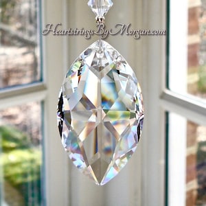 May include: A crystal suncatcher with a teardrop-shaped crystal hanging from a chain of smaller crystals. The suncatcher is hanging in front of a window with a view of trees and a brick wall. The text "Heartstrings By Morgan.com" is visible in the image.