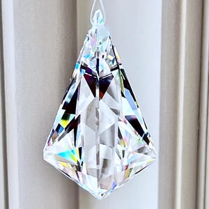 Puede incluir: Un colgante de prisma de cristal transparente y facetado cuelga de un hilo fino. El prisma tiene forma de diamante con m&uacute;ltiples superficies reflectantes, creando un efecto brillante. El sitio web HeartstringsByMorgan.com es visible en la parte inferior.
