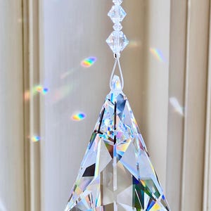 Puede incluir: Un atrapasoles de prisma de cristal transparente y facetado con un dise&ntilde;o geom&eacute;trico, suspendido por un fino hilo blanco. El prisma es triangular con una base en forma de diamante. La imagen muestra refracciones de luz arco&iacute;ris. Texto: Heartstrings By Morgan.