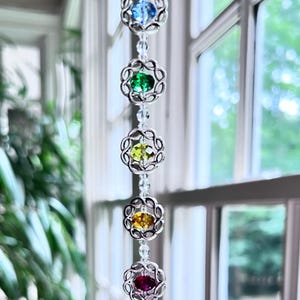 May include: A crystal suncatcher with a rainbow of colors hanging from a window. The suncatcher has a large crystal ball at the bottom and smaller crystals in a circle above it.