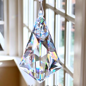 Puede incluir: Un atrapasol de prisma de cristal transparente y facetado, suspendido por un hilo fino. El prisma tiene forma de diamante con m&uacute;ltiples facetas, que reflejan un arco&iacute;ris de colores. El texto "Heartstrings By Morgan.com" es visible.