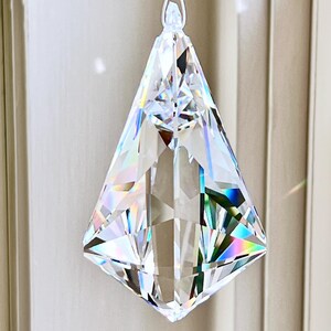 Puede incluir: Un colgante de prisma de cristal transparente y facetado con forma geom&eacute;trica, suspendido por una cadena de cuentas transparentes. El prisma refleja un arco&iacute;ris de colores. La direcci&oacute;n del sitio web Heartstrings By Morgan.com es visible.