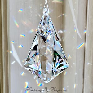 Puede incluir: Un prisma de cristal transparente y facetado suspendido de un hilo blanco. El prisma en forma de diamante tiene m&uacute;ltiples superficies reflectantes, creando patrones de luz de arco&iacute;ris en la pared. El texto "Heartstrings By Morgan.com" es visible.