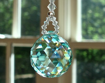 Op de afbeelding: Een gefacetteerde kristallen bol zonnevanger, hangend aan een draad van heldere kralen. De bol is een mix van blauw, groen en helder, die licht reflecteert. De achtergrond is een vage weergave van een raam en groen.
