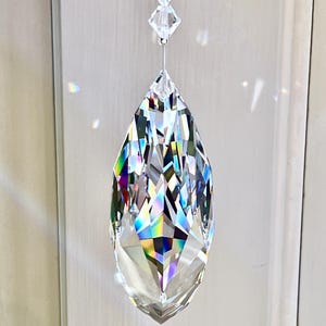 Puede incluir: Un gran colgante de prisma de cristal facetado, suspendido por un fino alambre y cristales m&aacute;s peque&ntilde;os. El colgante en forma de l&aacute;grima refleja un arco&iacute;ris de colores. El sitio web HeartstringsByMorgan.com es visible en la parte inferior.