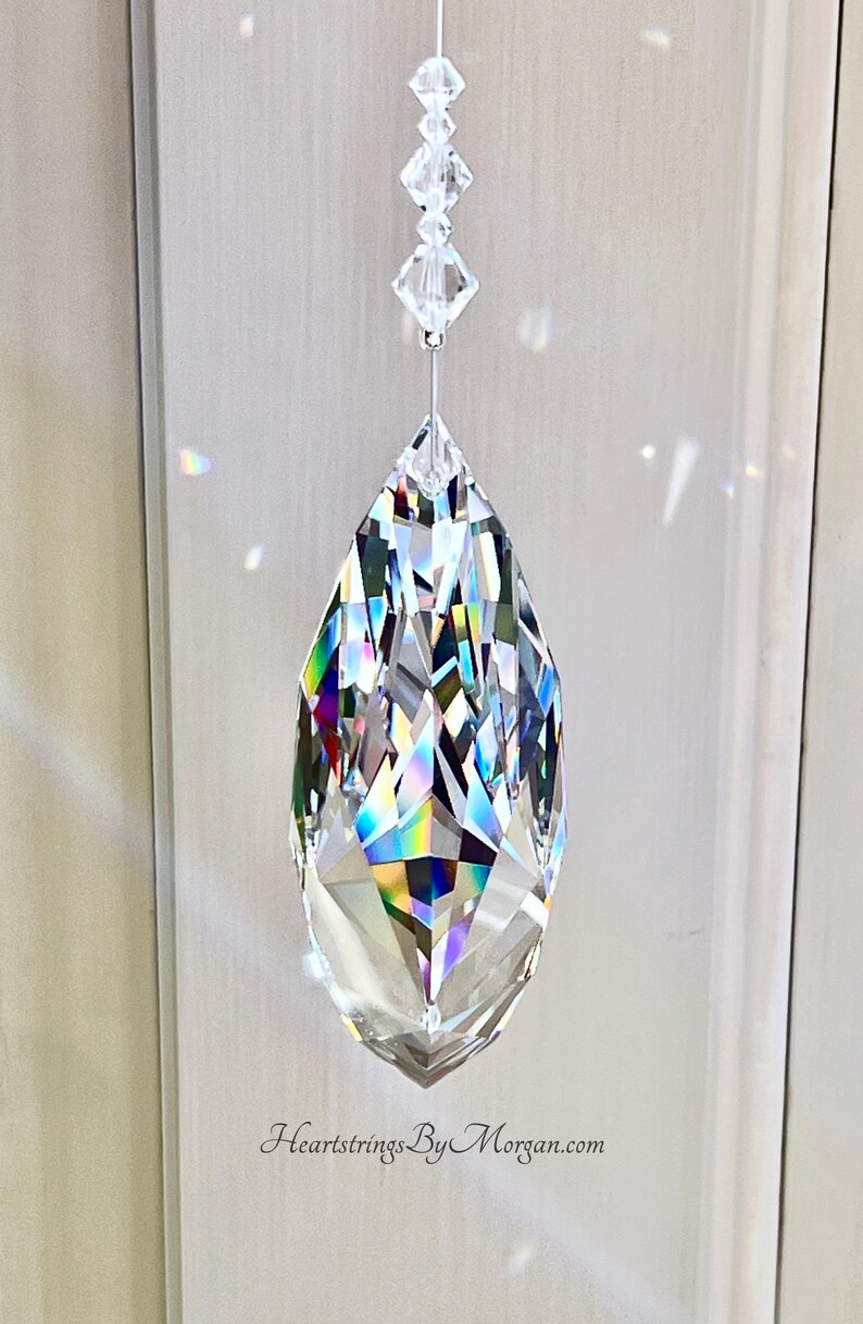 Puede incluir: Un gran colgante de prisma de cristal facetado, suspendido por un fino alambre y cristales m&aacute;s peque&ntilde;os. El colgante en forma de l&aacute;grima refleja un arco&iacute;ris de colores. El sitio web HeartstringsByMorgan.com es visible en la parte inferior.