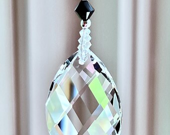 Op de afbeelding: Een kristallen zonenvanger met een gefacetteerde traanvorm hangt aan een ketting van zwarte en heldere kralen. De kristal is helder en reflecteert regenboogkleuren.