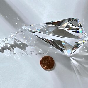 Puede incluir: Un gran prisma de cristal facetado con un extremo puntiagudo, suspendido por un alambre fino y peque&ntilde;as cuentas. El cristal refleja la luz, creando un efecto arco&iacute;ris. Una moneda est&aacute; al lado del cristal para la comparaci&oacute;n de tama&ntilde;os. El sitio web "HeartstringsByMorgan.com" es visible.