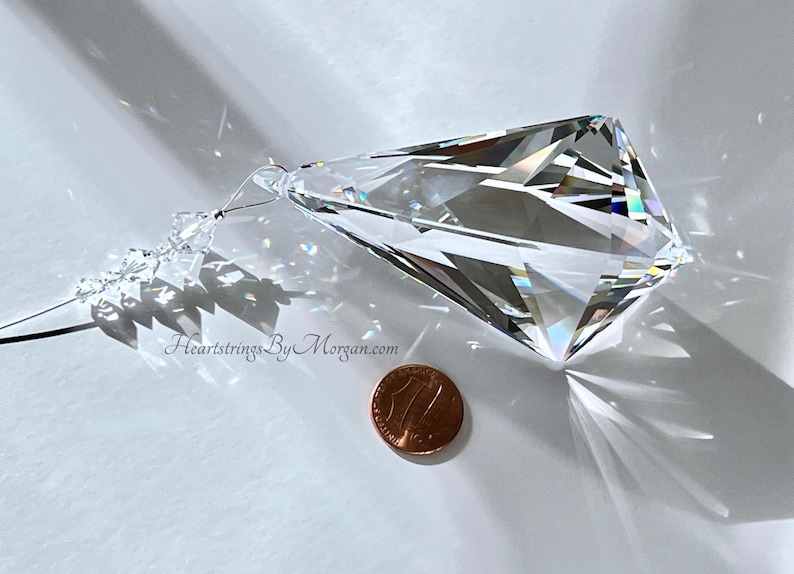 Puede incluir: Un gran prisma de cristal facetado con un extremo puntiagudo, suspendido por un alambre fino y peque&ntilde;as cuentas. El cristal refleja la luz, creando un efecto arco&iacute;ris. Una moneda est&aacute; al lado del cristal para la comparaci&oacute;n de tama&ntilde;os. El sitio web "HeartstringsByMorgan.com" es visible.