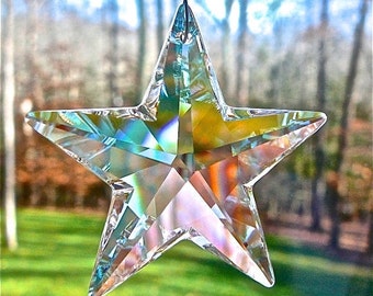 May include: A clear, faceted crystal star ornament. The star is suspended against a blurred background of green grass and trees. The crystal reflects light, displaying a rainbow of colours, including blue, green, yellow, and pink.