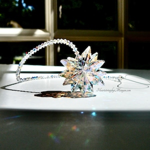 May include: A crystal star suncatcher with a clear iridescent finish hangs from a beaded chain. The star is casting rainbows on a white surface.